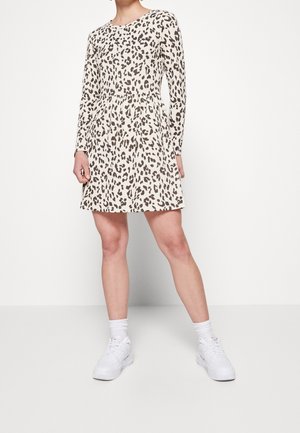 Vrouw die een lange mouw, boven-de-knie luipaardprint jurk draagt, witte enkelsokken en witte sneakers, staande tegen een effen witte achtergrond.