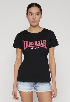 Jeune femme aux cheveux plaqués en arrière, portant un t-shirt noir "Lonsdale London" et un short en denim clair, sur un fond uni.