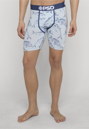 Mężczyzna w jasnoniebieskich bokserkach z ciemnoniebieskim wzorem ryb oraz granatową gumką PSD, stojący boso na neutralnym tle.