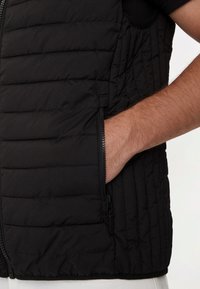 Gilet isolato nero con trapunte cucite, tasca con zip e design elegante. Il tessuto è morbido con una finitura opaca, hardware minimale.