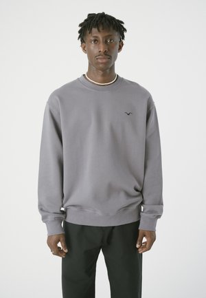 Grauer Rundhalsausschnitt-Pullover aus weichem Stoff mit gerippten Bündchen. Mit kleinem Logo auf der Brust. Kombiniert mit schwarzen Hosen.
