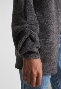 Pull en tricot gris avec une finition texturée. La manche présente un détail de poignet replié, associé à un jean en denim bleu.