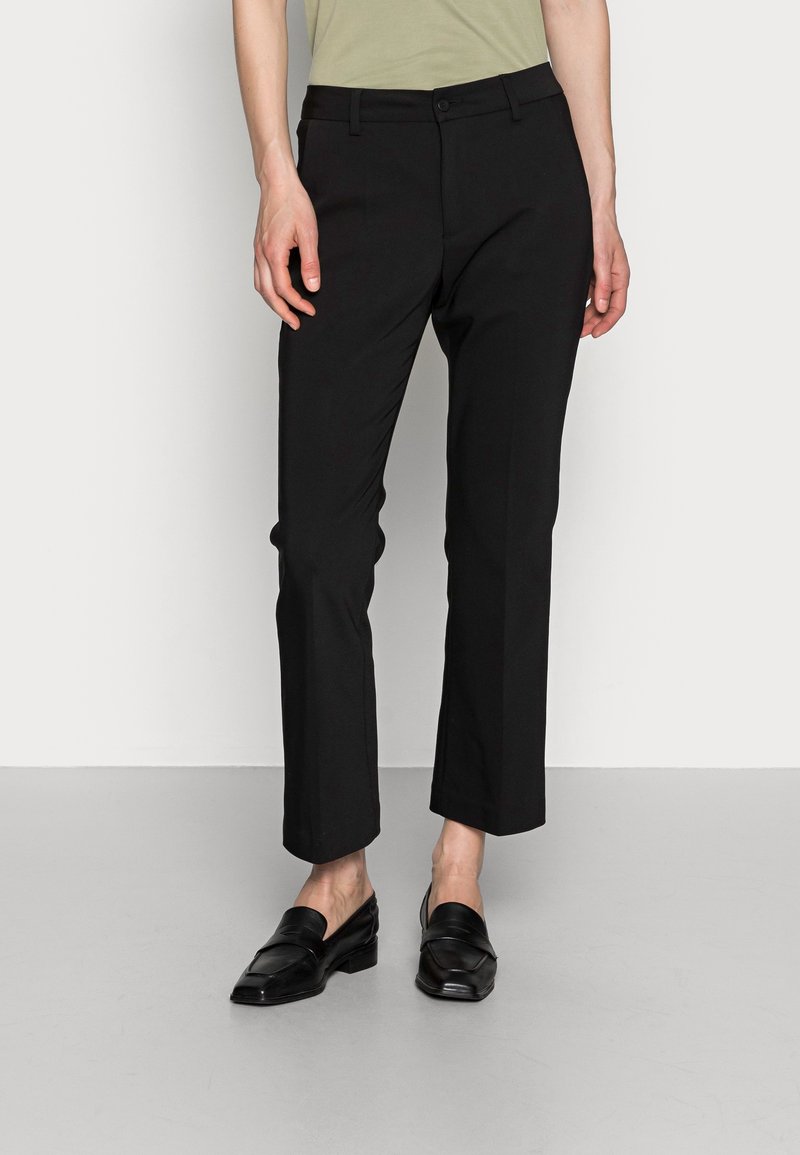 Personne portant un pantalon noir droit et des chaussures noires sans lacets, debout devant un fond clair uni.