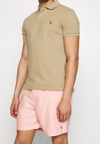 Polo shirt in cotone beige con un emblema verde sul petto, abbinato a pantaloni corti in lino rosa chiaro con un piccolo dettaglio del logo.