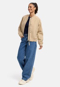 Veste bomber beige avec des poignets côtelés, associée à un jean en denim bleu ample. Des baskets blanches à semelle épaisse complètent la tenue.