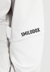 Witte sweatshirt van textuurstof; heeft een zwart 'SMILODOX' logo gedrukt op de rechtermouw. Slank fit met gestructureerde naden.