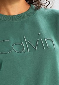 Zelená bavlněná mikina s žebrovaným límcem, zdobená vyšitým logem "Calvin" ve shodné barvě nitě. Měkká textilie a ležérní design.