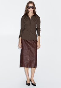 Braunes Hemd mit Knöpfen und Kragen, kombiniert mit einem braunen Leder-Midirock. Auffällige Sonnenbrillen und spitze schwarze Ballerinas runden den Look ab.