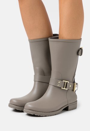 Botas de goma taupe hasta la rodilla con correas laterales y hebillas doradas, llevadas sobre piernas desnudas, presentadas sobre un fondo blanco sencillo.