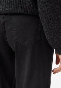 Zbliżenie na osobę w czarnych spodniach z tylną kieszenią i czarnym, teksturowanym swetrze, z ręką swobodnie opuszczoną wzdłuż ciała.