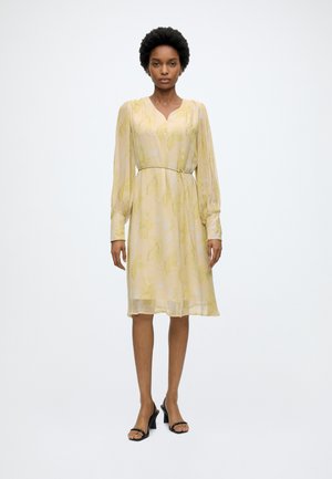 Γυναίκα που φοράει ένα ανοιχτό κίτρινο, μακρυμάνικο, φόρεμα μέχρι τα γόνατα με διακριτικά λουλουδάτα σχέδια, στέκεται μπροστά σε λευκό φόντο.