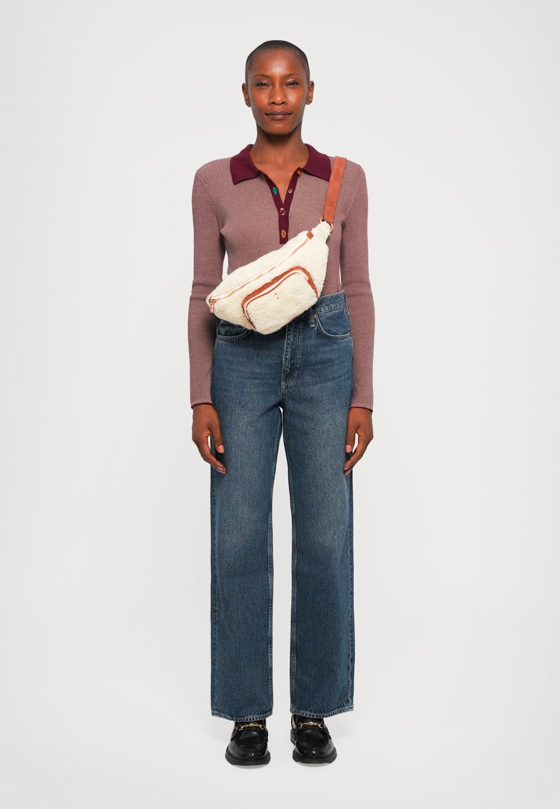 Beige Umhängetasche mit orangefarbenen Akzenten, weiche Textur; langärmliges lila Oberteil mit bordeauxfarbenem Kragen; weit geschnittene blaue Jeans; schwarze Schuhe.