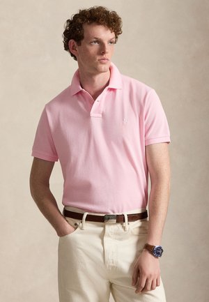 Młody mężczyzna o kręconych włosach, ubrany w różową koszulkę polo z krótkim rękawem, kremowe spodnie, brązowy pasek i niebieski zegarek, stojący z jedną ręką w kieszeni.