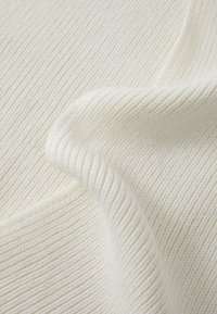 Detailný záber na jemnú ribbed pleteninu v krémovej farbe s jemnými záhybmi a detailmi textúry.