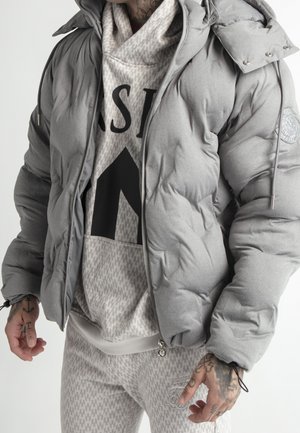 Mand iført en grå dunjakke over en mønstret beige hættetrøje og matchende bukser, der viser tatoverede hænder og en afslappet streetwear-stil.