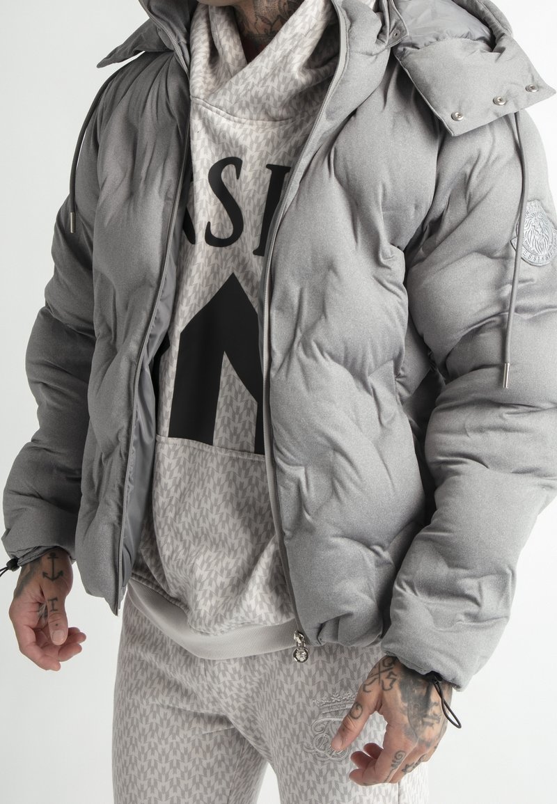 Homme portant une doudoune grise par-dessus un sweat à capuche beige à motifs et un pantalon assorti, montrant ses mains tatouées et un style décontracté de rue.