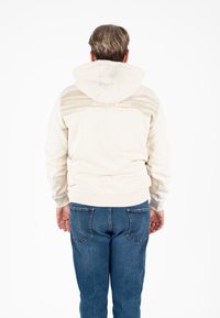 Lichtbeige hoodie met een grote capuchon, horizontaal gestreept ontwerp op de achterkant en geribde manchetten. Gecombineerd met blauwe jeans.