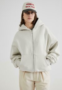 Felpa grigia chiara con zip in tessuto morbido, dotata di un grande cappuccio e tasche frontali. Abbinata a pantaloni beige e un cappellino con testo rosso.
