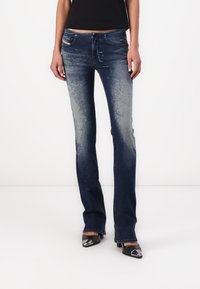 Jeans de tiro alto de color azul oscuro con patrones claros de desgaste y desvanecimiento. Presentan cinco bolsillos y un ajuste ceñido, combinados con zapatos negros de punta.