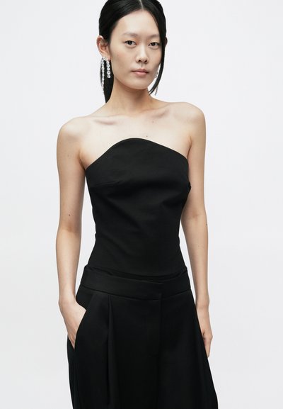 Melna strapless top ar strukturētu dizainu, gludu faktūru un piegulošu siluetu, kombinēts ar augsta vidukļa melnām biksēm ar kloķiem.