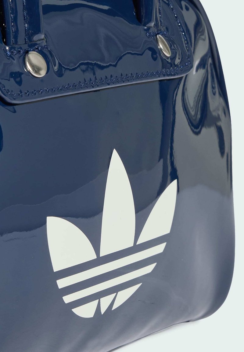 Glänzende dunkelblaue Tasche mit weißem, stilisiertem dreiblättrigem Logo und sichtbaren Metallnieten nahe der Griffbefestigung.
