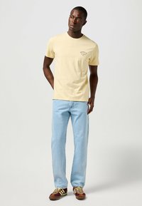 Camiseta de manga corta en color amarillo claro con un pequeño gráfico en el lado izquierdo del pecho, combinada con jeans claros y zapatillas marrones y amarillas.