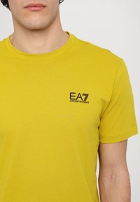 Sárga pamut póló kerek nyakkal, rövid ujjal, és egy kis fekete EA7 logóval a bal mellrészen.