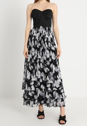 Femme portant une robe noire bustier texturée avec une jupe florale noire et blanche à plusieurs couches, associée à des escarpins ouverts noirs.