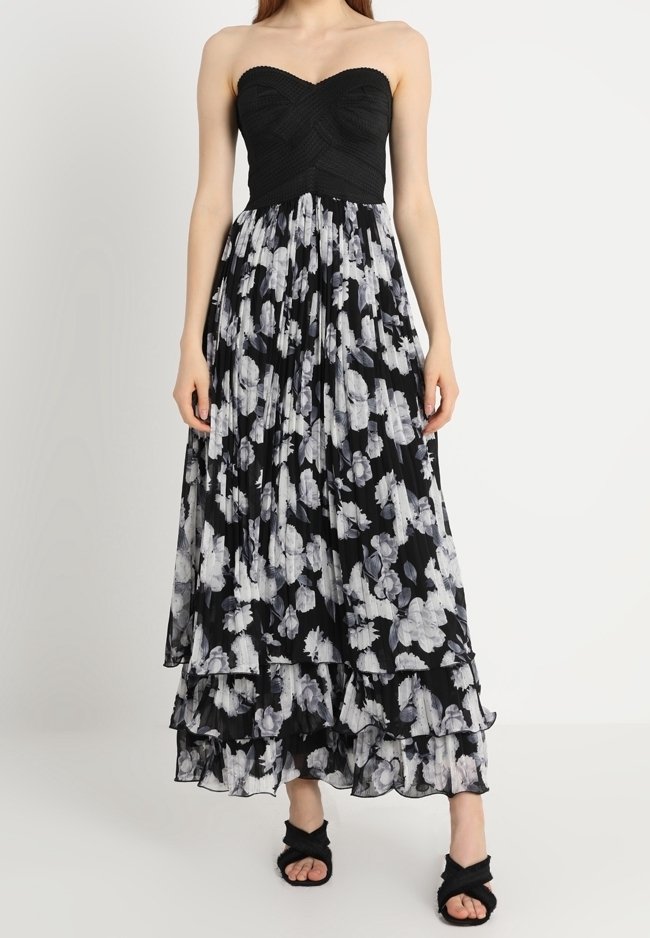 Femme portant une robe noire bustier texturée avec une jupe florale noire et blanche à plusieurs couches, associée à des escarpins ouverts noirs.