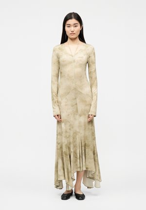 Femme debout de face, portant une robe beige à manches longues avec un motif subtil et un ourlet asymétrique, assortie de chaussures plates noires.