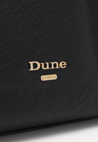 Černá kožená taška s zlatým logem "Dune" a malým zlatým akcentem "LONDON", s texturovaným povrchem a strukturovaným designem.