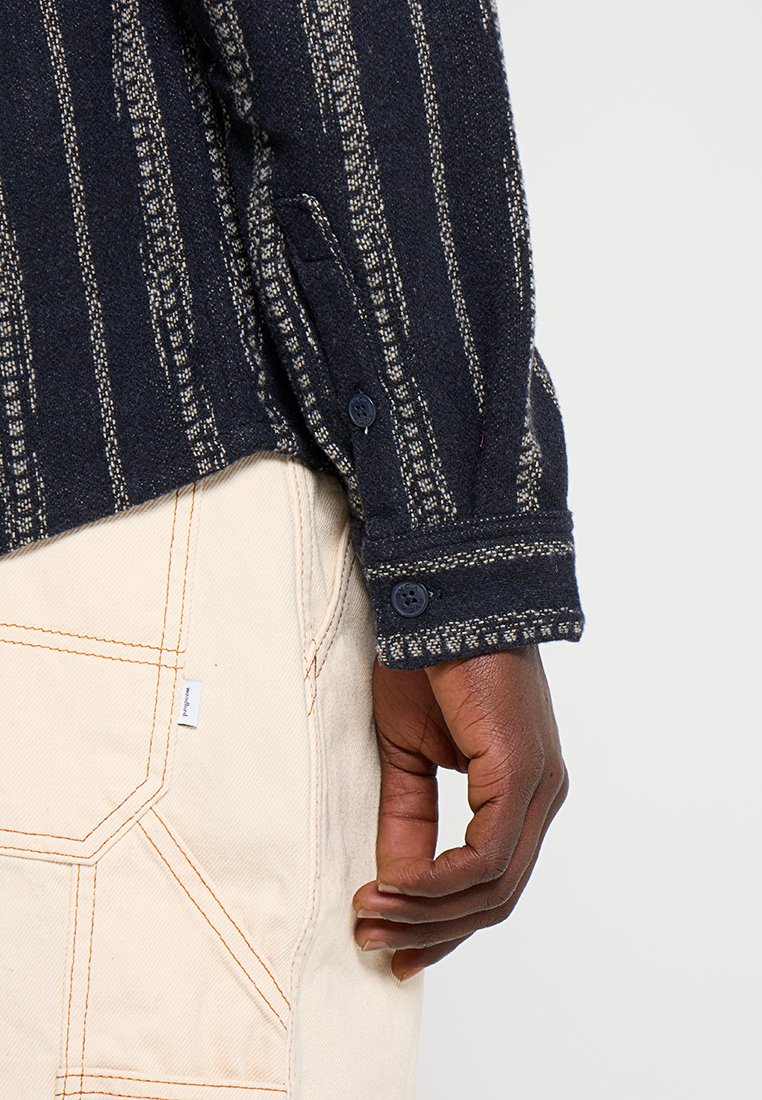 Marineblauwe gestreepte shirt met een textuurstof, voorzien van knopenmanchetten en witte accentlijnen, gecombineerd met beige broek met zichtbare stiksels.