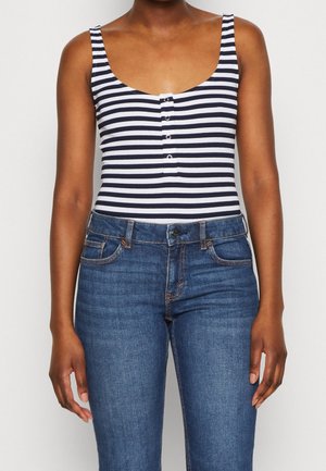 Persona vistiendo una blusa sin mangas de rayas blancas y negras con botones, combinada con jeans de mezclilla azul sobre un fondo liso.