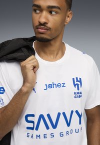 Egy férfi fehér sportmezben, kék logókkal és felirattal, feketére színezett dzsekit tart a válla fölött, balra nézve.