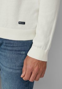 Heller Cremepullover mit Rippbündchen an Ärmeln und Saum, aus weichem Stoff, mit einem kleinen schwarzen Etikett an der Seite, kombiniert mit blauer Jeans.