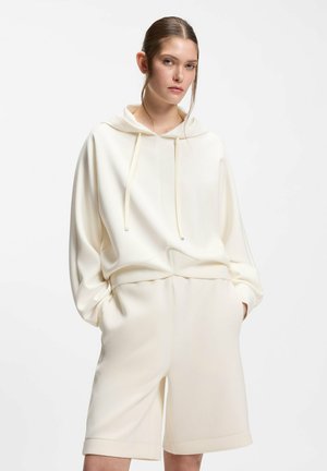 Jonge vrouw met bruin haar naar achteren gebonden, draagt een oversized crème hoodie en bijpassende knielange shorts met handen in de zakken.