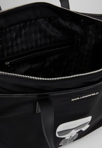 Svart tygväska med dragkedjestängning, med ett inre foder med ett subtilt mönster, en dragkedjeficka och en vit grafisk logotyp på framsidan.