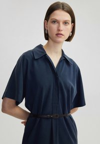 Abito blu navy a maniche corte con colletto, caratterizzato da un'apertura frontale con bottoni e una cintura nera arricciata in vita. Tessuto liscio e texture.