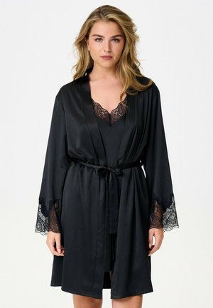 Jeune femme aux cheveux blonds portant une robe en satin noir avec un bord en dentelle aux manches et à l'encolure, nouée à la taille avec une ceinture.