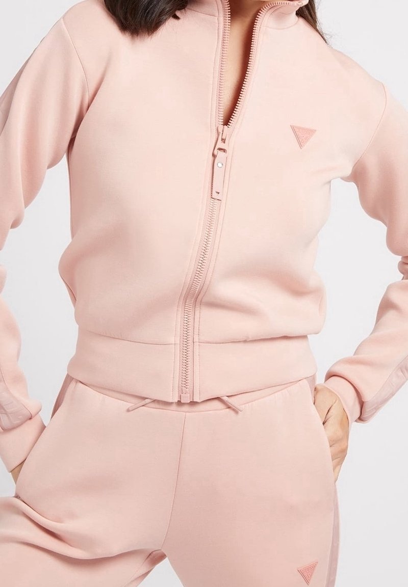 Růžová zipová bunda s odpovídajícími kalhotami, vyrobená z měkké látky. Vlastnosti zahrnují vysoký límec, boční kapsy a trojúhelníkové logo na hrudi.