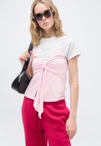 Νεαρή γυναίκα που φοράει ροζ τοπ με στρώσεις, φωτεινά ροζ παντελόνια, μαύρη τσάντα ώμου και σκούρα γυαλιά ηλίου, πάνω σε ένα μονόχρωμο φόντο.