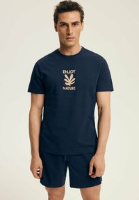 Granatowa bawełniana koszulka z jasnobeżowym nadrukiem z napisem "CIESZ SIĘ PRZYRODĄ" oraz wzorem liścia; noszona z pasującymi szortami.