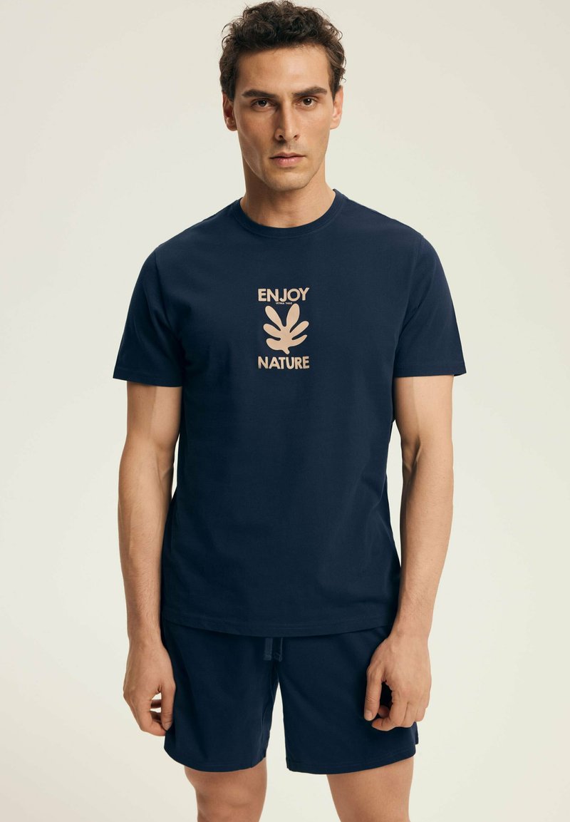 Granatowa bawełniana koszulka z jasnobeżowym nadrukiem z napisem "CIESZ SIĘ PRZYRODĄ" oraz wzorem liścia; noszona z pasującymi szortami.