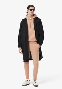 Giacca leggera nera sopra un hoodie pesca e pantaloni tuta abbinati. Il modello indossa occhiali da sole e sneakers bianche con suola marrone.