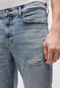 Jeans de mezclilla azul claro con detalle desgarbado en el muslo, cinco bolsillos y costuras visibles a lo largo de las costuras.