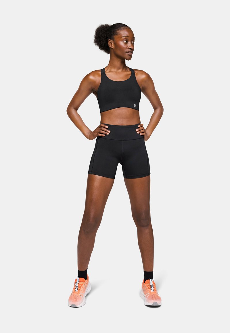 Sujetador deportivo negro y pantalones cortos, ambos de tejido elástico. Las zapatillas son de color rosa claro con detalles en negro. El modelo posa con las manos en las caderas.