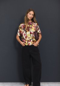 Farebné batikované tričko z mäkkej látky s krátkymi rukávmi, v kombinácii s čiernymi širokými nohavicami. V ruke drží tmavú kabelku.