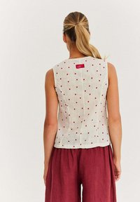 Bezrękawnik z lnu w białym kolorze w czerwone kropki, z metką z tyłu i dopasowanym fasonem; zestawiony z luźnymi burgundowymi spodniami.