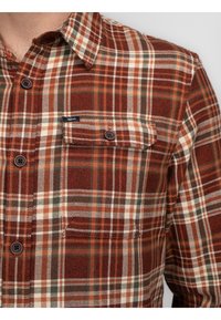 Camisa de franela a cuadros en marrón y crema con un bolsillo en el pecho, botones oscuros y acentos verdes sutiles. Textura suave y diseño casual.