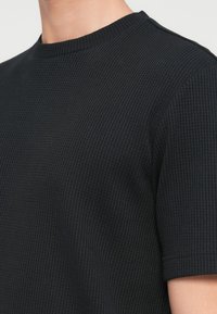 Μαύρο κοντομάνικο πουλόβερ με υφή πλεξίματος και στρογγυλή λαιμόκοψη, φορεμένο από άτομο και φαίνεται από τον ώμο μέχρι το μέσο του στήθους.